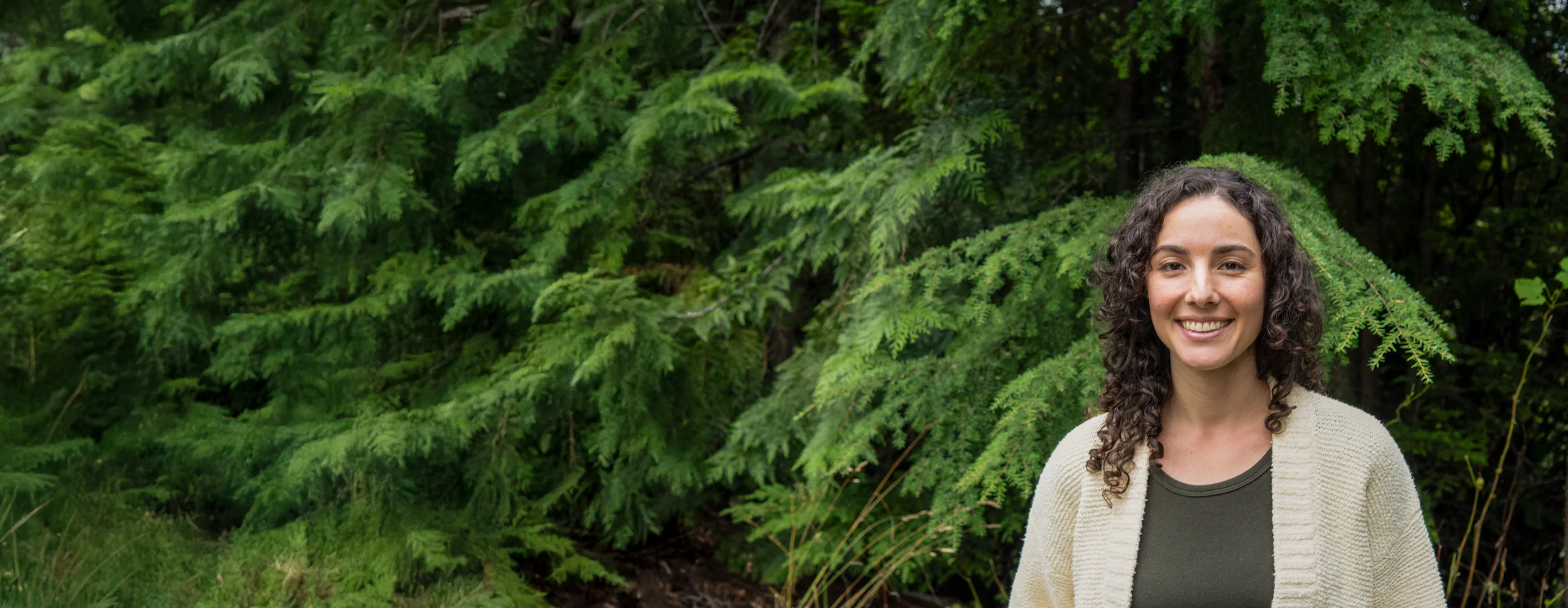 Vanessa Deverell smiling, in a cream cardigan, with cedar branches behind her, near Nelson, BC.