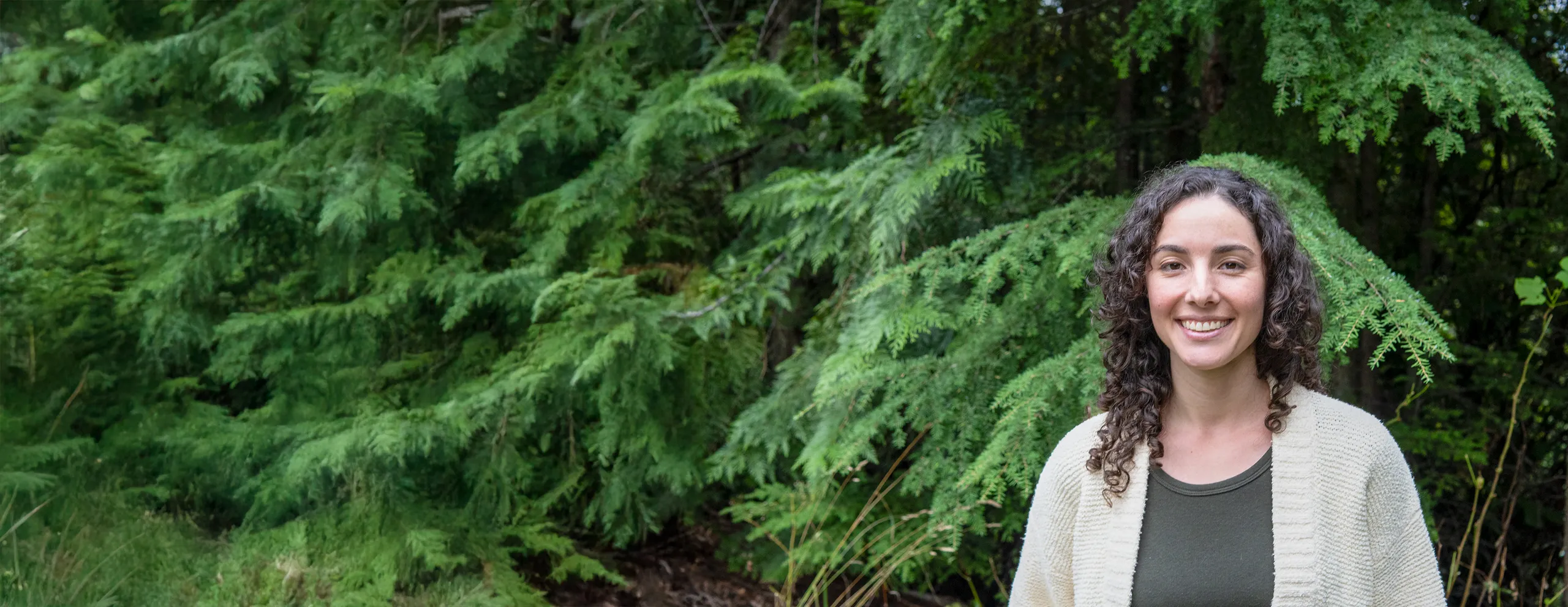 Vanessa Deverell smiling, in a cream cardigan, with cedar branches behind her, near Nelson, BC.